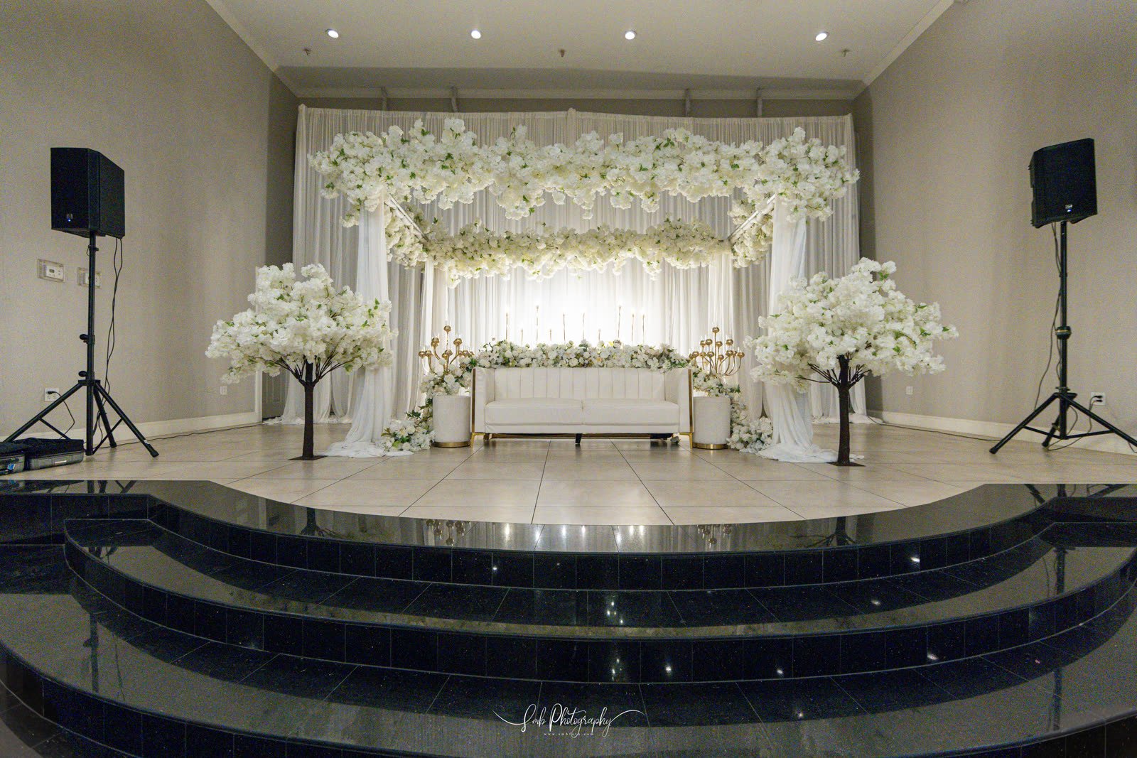 A ceremony stage styled with white florals and blossom trees at Marquee